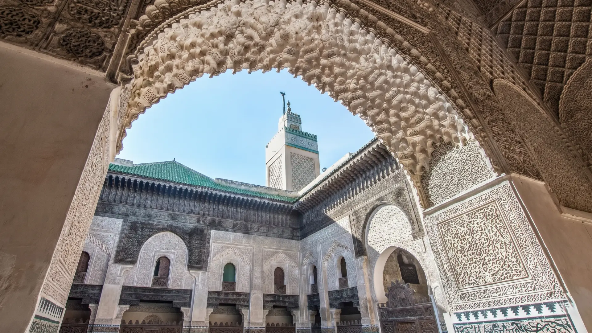 Detailed stone carved arch framing a green-tiled minaret and courtyard in historic Fez medersa Morocco