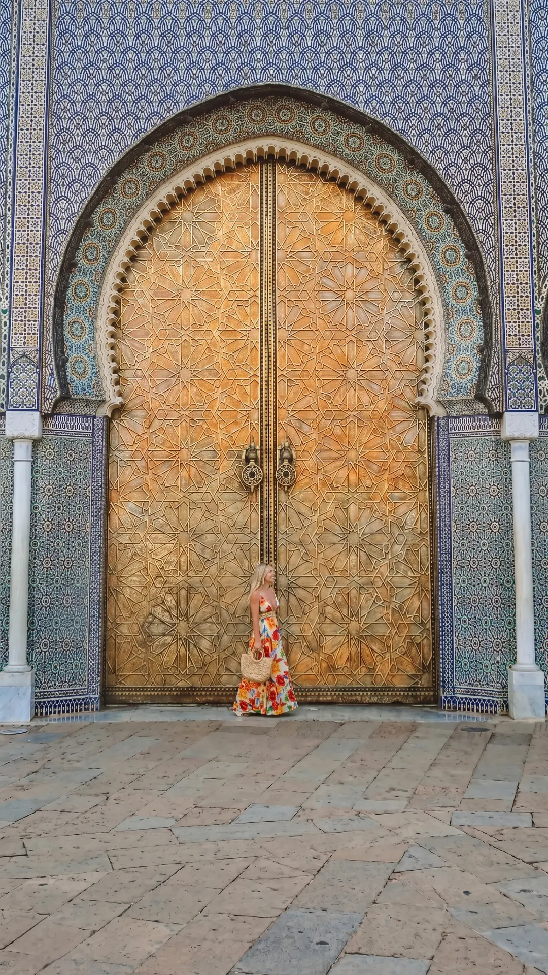 Woman standing beside enormous intricately carved golden wooden doors with blue zellige tiles in Fez